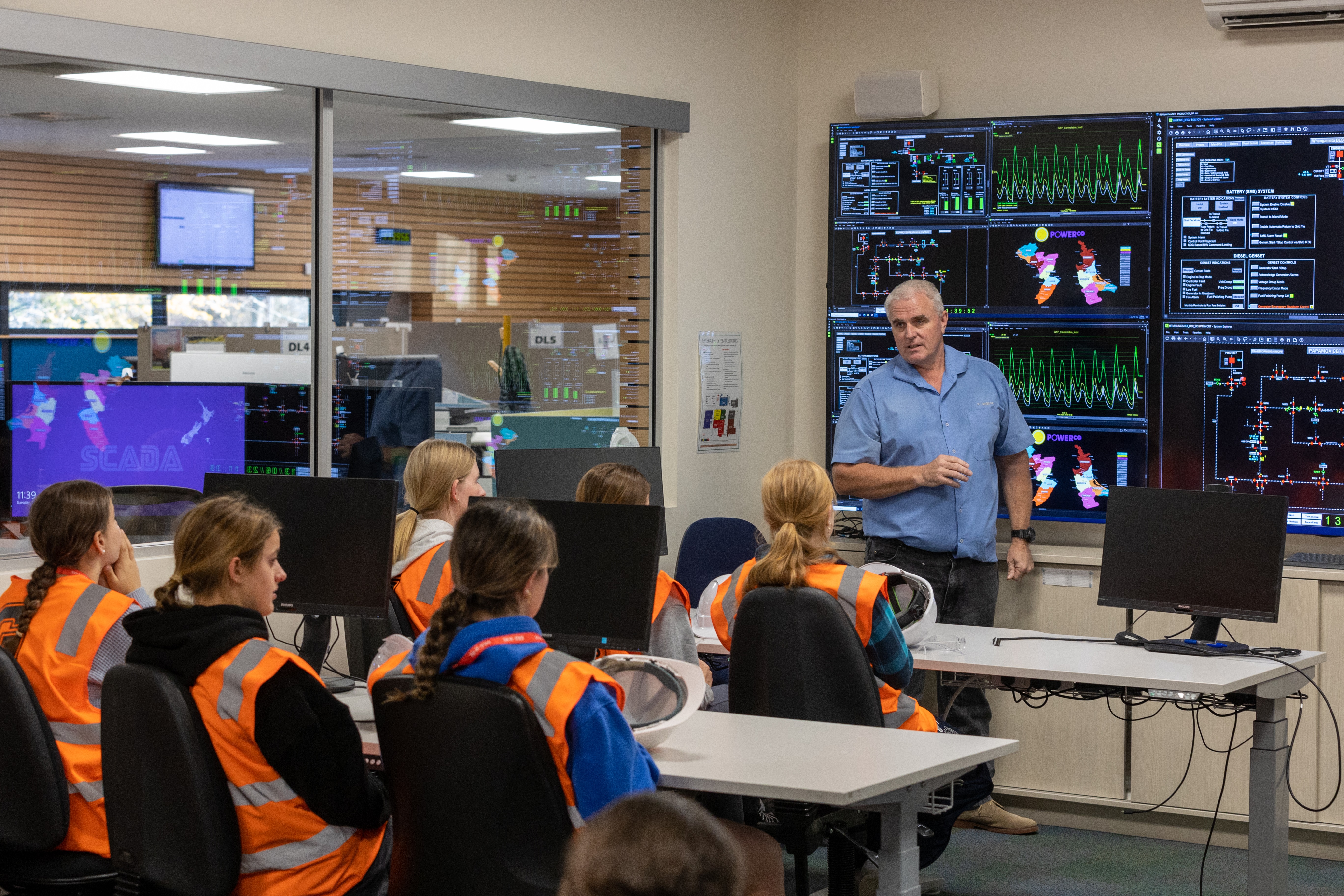 A man with Scada screens behind him, teaching a group of girls wearing hi-vis about the electricity distribution industry