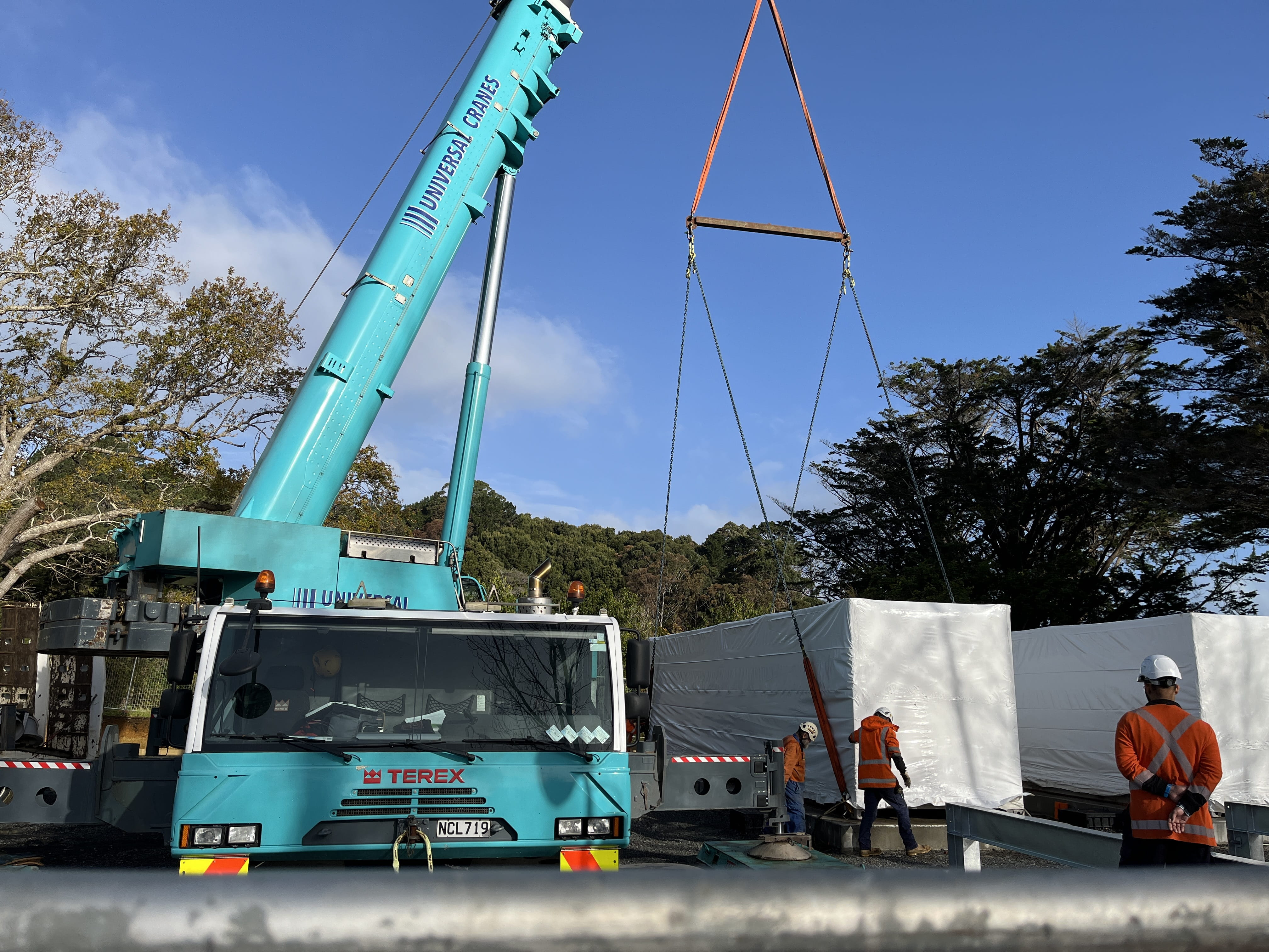 A shipping container sized generator, wrapped in plastic, is lowered to the ground by a crane