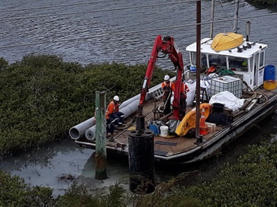 A barge on the side of an estuary with a digger on it and workmen erecting a power poles