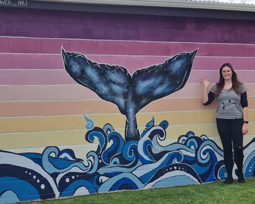A woman next to a tropical coloured mural featuring a whale tail coming out of waves.