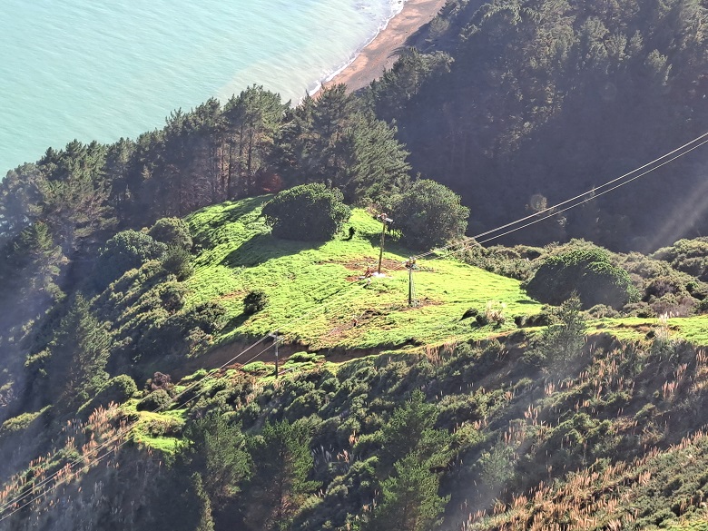 Helicopter view of remote, green hill with trees and sea and electricity crew working on power poles