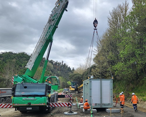 A green crane in a rural area lowering a generator container onto a concrete platform with workers in PPE